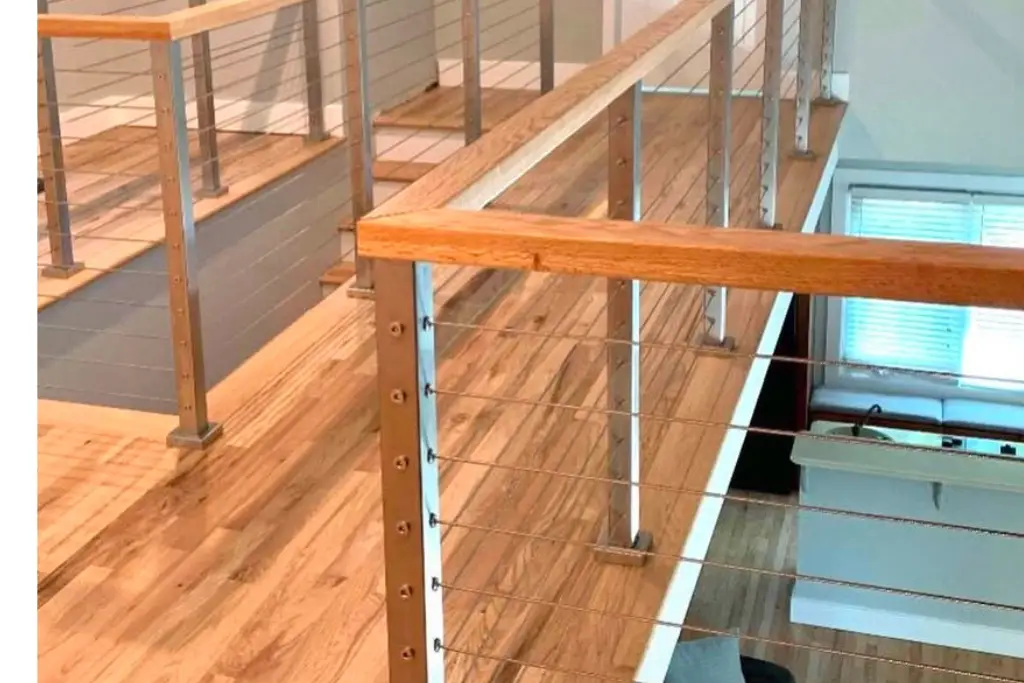 “Interior cable railing with silver stainless steel posts and a brown wooden handrail, installed along a staircase in a modern home.